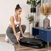 A woman is kneeling on the floor next to a black vibration plate in a living room. Voir les images du produit