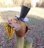 A decorative metal bird sculpture with a top hat and umbrella, mounted on a wooden post in an outdoor setting with fallen leaves. Produktbild-Vorschau