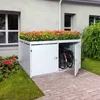 A white bicycle storage shed with a green roof covered in colorful flowers, situated next to a house and on a paved walkway. Náhled obrázku produktu