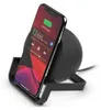 A black wireless charging stand with a smartphone placed on it, showing the phone's screen with music playing and battery at 100%. Produktbild-Vorschau
