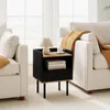 A black and wood-tone nightstand with a lamp, a cup, and books on it, placed between two cream-colored sofas in a well-lit room. Náhled obrázku produktu