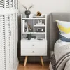 A white nightstand with two drawers and shelves is placed next to a bed in a bedroom. Podgląd zdjęcia produktu