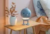 A blue robot-shaped lamp sits on a wooden side table next to a vase with dried leaves and a bed with an orange blanket. Produktbild-Vorschau