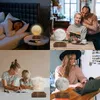 A collage of four images showing a levitating moon lamp in different settings: a bedroom, a living room with a couple reading, and a table with children reading. Another image shows a woman working on a laptop with the lamp nearby. Náhled obrázku produktu