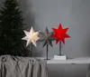 Three star-shaped lamps are displayed on a table next to a Christmas tree. One lamp is beige, one is gray, and one is red. A gray tablecloth is draped over the table in the foreground. Podgląd zdjęcia produktu