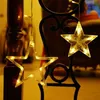 Close-up of two illuminated star-shaped string lights hanging from a dark metal structure, with a blurred wooden pillar in the background. Podgląd zdjęcia produktu