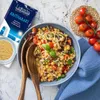A bowl of orzo pasta salad with chickpeas, tomatoes, and olives, next to a package of Kritharaki pasta and a bunch of cherry tomatoes. Podgląd zdjęcia produktu