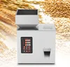 A white automated particle weighing and filling machine is shown in the center, with a hopper on top and a control panel on the front. The background features blurred images of wheat and oats. Náhľad obrázku produktu