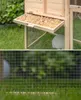 A split image showing the top half of a wooden rabbit hutch with a pull-out tray filled with wood shavings, and the bottom half showing a wire mesh fence in front of green grass and blurred foliage. Produktbild-Vorschau