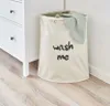 A beige laundry hamper with the words "wash me" printed on it, placed next to a white dresser. A green towel is draped over the edge of the hamper. Produktbild-Vorschau