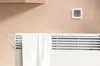 A digital thermometer is mounted on a wall above a white radiator with a towel draped over it. Podgląd zdjęcia produktu