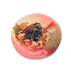 A bee is shown on a pile of seeds inside a round, pink and yellow container. Podgląd zdjęcia produktu