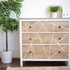 A dresser with a wood-look finish and geometric white lines on the drawers, next to a potted plant. Produktbild-Vorschau
