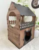 A wooden cat house with two levels, featuring a cat-shaped entrance on the lower level and a balcony with a fence on the upper level. It is placed on a white, textured surface with blurred white flowers and a clock in the background. Voir les images du produit