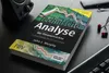 A book titled 'Technische Analyse der Finanzmärkte' is placed on a dark wooden surface, surrounded by a speaker, earphones, and a keyboard. Náhľad obrázku produktu