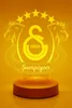 A 3D LED lamp with the Galatasaray logo and the word 'Şampiyon' (Champion) on a wooden base, illuminated with yellow light against an orange background. Produktbild-Vorschau