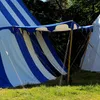 A blue and white striped tent with a canopy supported by wooden poles, set up on a grassy field with trees in the background. Produktbild-Vorschau