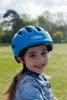 A young girl wearing a blue bicycle helmet smiles at the camera in an outdoor setting. Produktbild-Vorschau