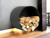 A black, circular metal log holder mounted on a gray wall, filled with firewood. The holder is positioned above a wooden floor with a strip of light running along the bottom edge of the wall. Produktbild-Vorschau