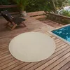 A round beige rug is placed on a wooden deck next to a swimming pool. There are lounge chairs and a bench on the deck, with trees and water in the background. Náhled obrázku produktu