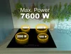 A black induction cooktop with four illuminated cooking zones, each displaying a power output. The background features a backsplash with dried herbs and a countertop with kitchen accessories. Produktbild-Vorschau