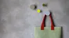 A light green tote bag with red handles hangs from a gray circular hook on a textured gray wall. Three other circular hooks in gray, yellow, and olive green are also visible on the wall. Produktbild-Vorschau