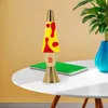 A lava lamp sits on a white table next to a green notebook and a blue vase with a plant. Anteprima dell’immagine prodotto