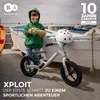 A young child in a green hoodie is holding a toy near a balance bike with a white helmet on its handlebars, with boats in the background. Produktbild-Vorschau