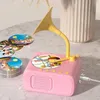 A pink toy record player with a yellow horn sits on a textured surface, with a stack of colorful discs next to it. A USB cable is plugged into the side. Produktbild-Vorschau
