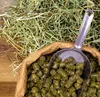 A close-up view of deukavallo Heucobs in a brown paper bag, with a metal scoop filled with the pellets. A pile of hay is visible in the background. Produktbild-Vorschau