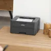 A black Brother laser printer is shown on a wooden surface, with a piece of paper coming out of it. There are two wooden candle holders to the right of the printer. Produktbild-Vorschau