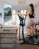A woman is using an elliptical cross-trainer in a room that appears to be a living room or bedroom, with a large screen displaying a scenic road. Produktbild-Vorschau