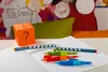 A collection of colorful pens and pencils are arranged on an open notebook, with a question mark cube in the background. The scene is set on a desk with children's artwork in the background. Anteprima dell’immagine prodotto