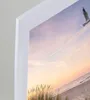 A close-up view of a white picture frame with a beach scene at sunset visible inside. Produktbild-Vorschau