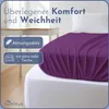A close-up of a purple fitted sheet draped over a white mattress, with text overlays highlighting product features. Produktbild-Vorschau