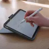 A person's hand is using a stylus to write on a tablet, which is placed on a wooden table. Náhled obrázku produktu