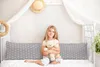 A young girl sits on a bed holding a teddy bear, with a large pillow behind her. Produktbild-Vorschau