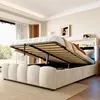A bedroom with a bed that has its storage compartment open, revealing the mattress and frame. The room has a large window with curtains, a picture on the wall, and a bedside table with a tablet. Náhľad obrázku produktu