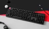 A black mechanical keyboard is laid out on a surface with black, red, and gray sections. There are also some small spherical objects and pencils in the background. Produktbild-Vorschau