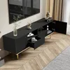A black TV stand with gold legs is shown in a living room setting, with a TV mounted on the wall above it. The stand has open drawers and doors, revealing some items inside. Curtains are visible in the background. Produktbild-Vorschau