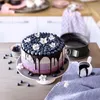 A decorated cake with blueberries and flowers on a table, with baking equipment in the background. Náhled obrázku produktu