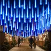 A street scene at night decorated with blue meteor shower lights, with people walking by Christmas trees and market stalls. Anteprima dell’immagine prodotto