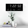 A digital clock with calendar and temperature display mounted on a white wall above a table with decorative items. Produktbild-Vorschau