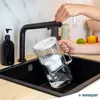 A person is filling a glass water filter pitcher under a black faucet in a kitchen sink. Náhľad obrázku produktu
