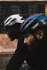 Two men wearing cycling helmets, one white and one iridescent blue, are shown in profile, riding bicycles. Podgląd zdjęcia produktu