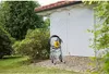 A yellow and gray airless paint sprayer is set up on a brick patio in front of a white garage door, surrounded by lush green trees and grass. Náhľad obrázku produktu