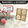 Christmas decorations, including straw stars and a red bauble, are arranged on a surface with a blurred background of Christmas lights and pine branches. Produktbild-Vorschau