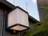 A modern, cube-shaped outdoor wall light with a motion sensor is mounted on a wooden fence, with green foliage in the foreground and a blurred background of a building and sky. Anteprima dell’immagine prodotto