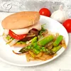 A close-up of a burger and asparagus on a white plate, with tomatoes and garlic in the background. Produktbild-Vorschau