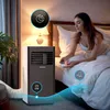 A woman is sitting on a bed, holding a remote control, and looking at a portable air conditioner in a bedroom. Produktbild-Vorschau
